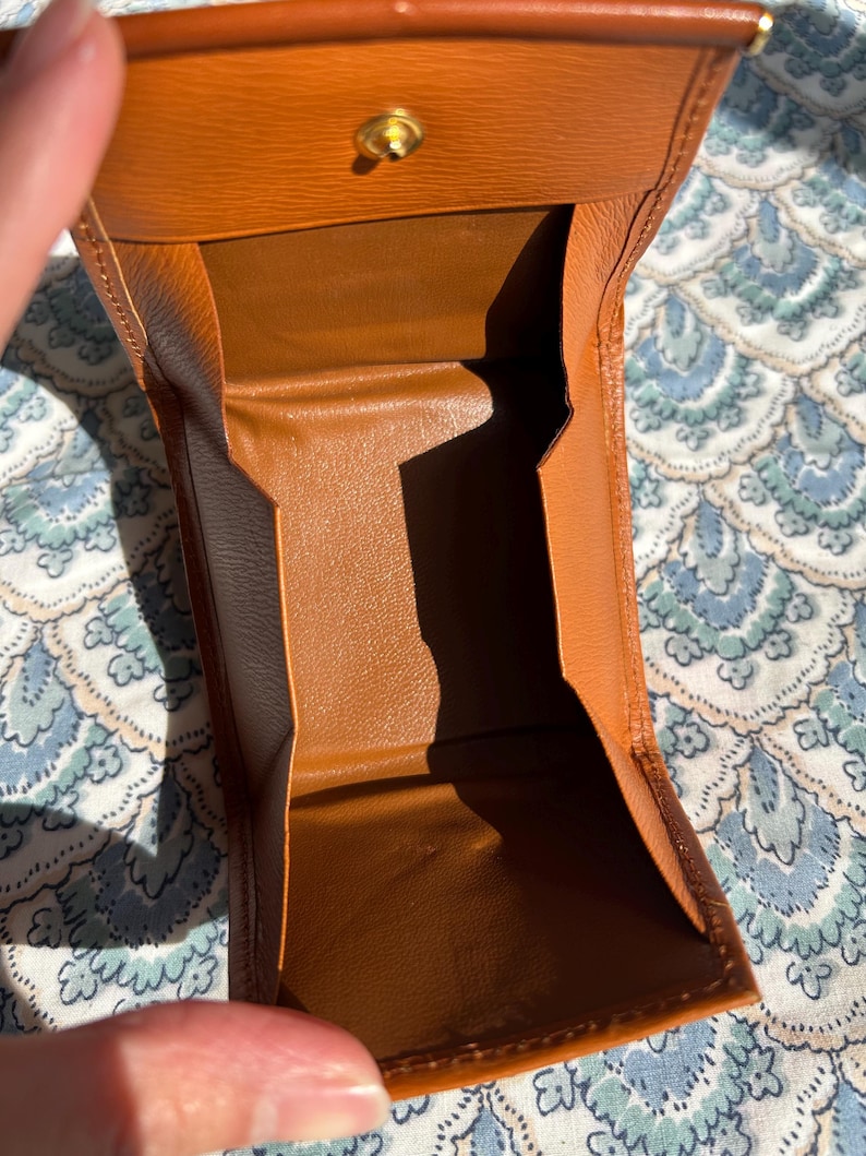 May include: Open, brown leather wallet with a gold-colored clasp. The wallet is open, revealing the interior compartments. The leather has a textured appearance. The wallet is on a patterned surface.