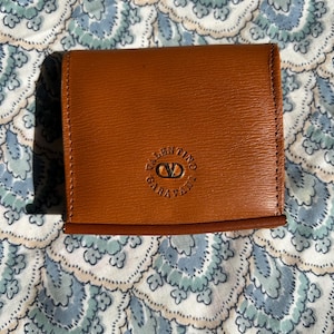 May include: A brown leather coin purse with the Valentino Garavani logo embossed on the front. The purse is rectangular and has a stitched border. The background is a blue and white floral pattern.