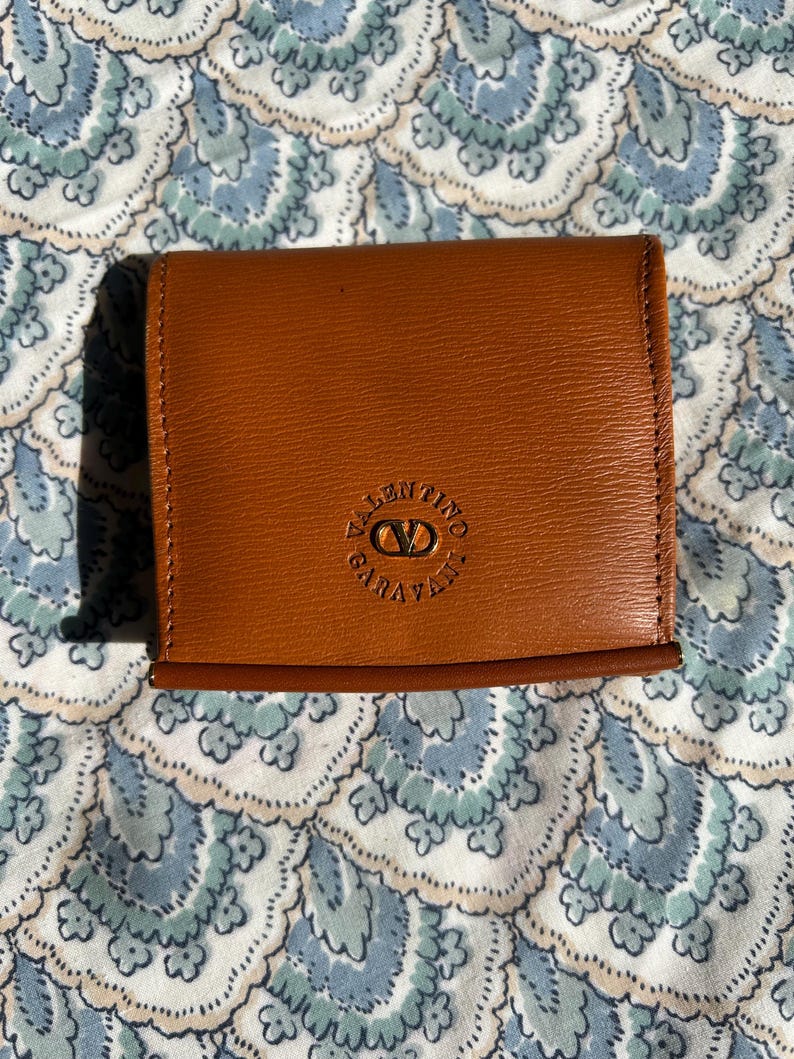 May include: A brown leather coin purse with the Valentino Garavani logo embossed on the front. The purse is rectangular and has a stitched border. The background is a blue and white floral pattern.