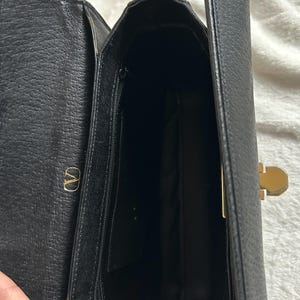 May include: Open view of a black leather handbag with a gold-colored clasp and a zipper pocket. The interior is visible, revealing a spacious compartment. The bag features a gold "V" logo on the side.