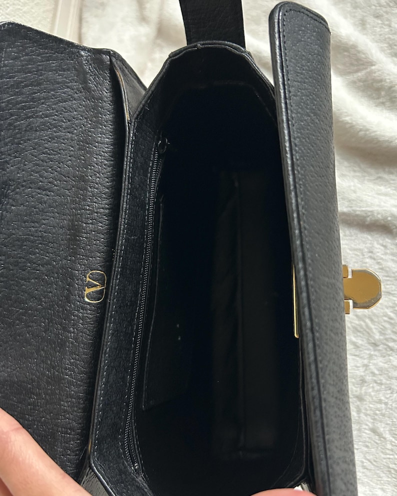 May include: Open view of a black leather handbag with a gold-colored clasp and a zipper pocket. The interior is visible, revealing a spacious compartment. The bag features a gold "V" logo on the side.