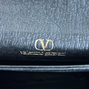 May include: Close-up of a black leather handbag featuring a gold-colored "V" logo and the brand name "Valentino Garavani". The bag has a textured surface.
