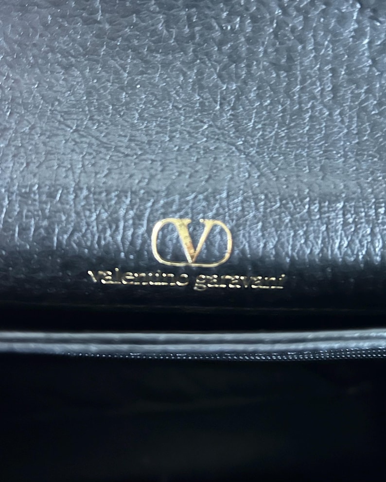 May include: Close-up of a black leather handbag featuring a gold-colored "V" logo and the brand name "Valentino Garavani". The bag has a textured surface.