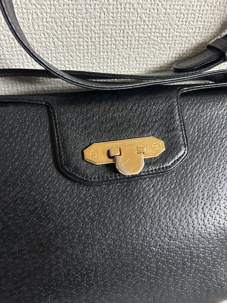 May include: Black leather handbag with a gold-tone clasp. The handbag features a textured surface and a shoulder strap. The clasp is rectangular with the words "valentino garavani" engraved on it.
