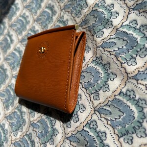 May include: A small, brown leather wallet with a gold-colored clasp and a decorative emblem. The wallet is rectangular and appears to be new. The background is a patterned fabric in shades of blue and white.