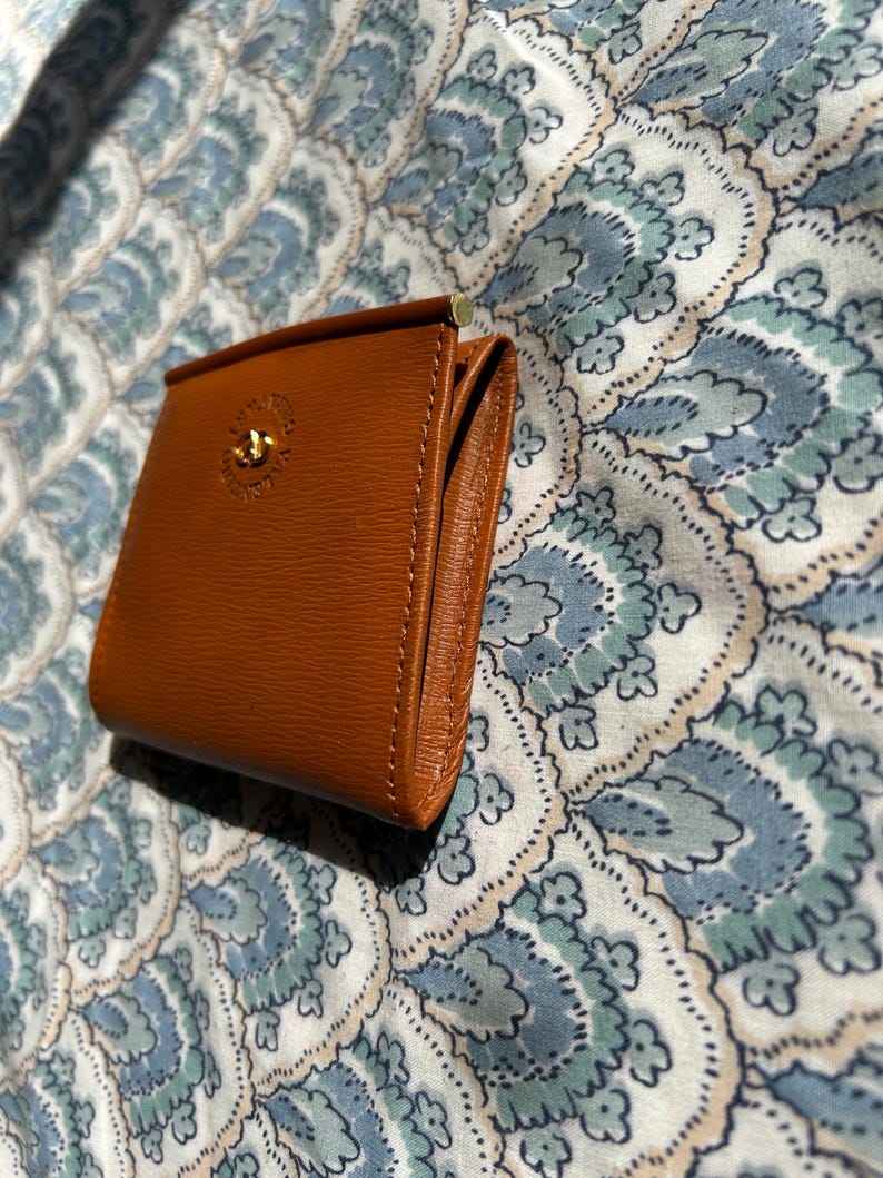 May include: A small, brown leather wallet with a gold-colored clasp and a decorative emblem. The wallet is rectangular and appears to be new. The background is a patterned fabric in shades of blue and white.