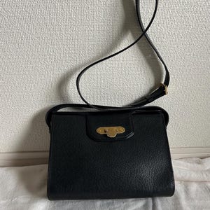 May include: Black leather shoulder bag with a rectangular shape and a long, thin strap. The bag features a gold-colored clasp on the front and a textured surface. The strap is adjustable and appears to be made of the same material as the bag.
