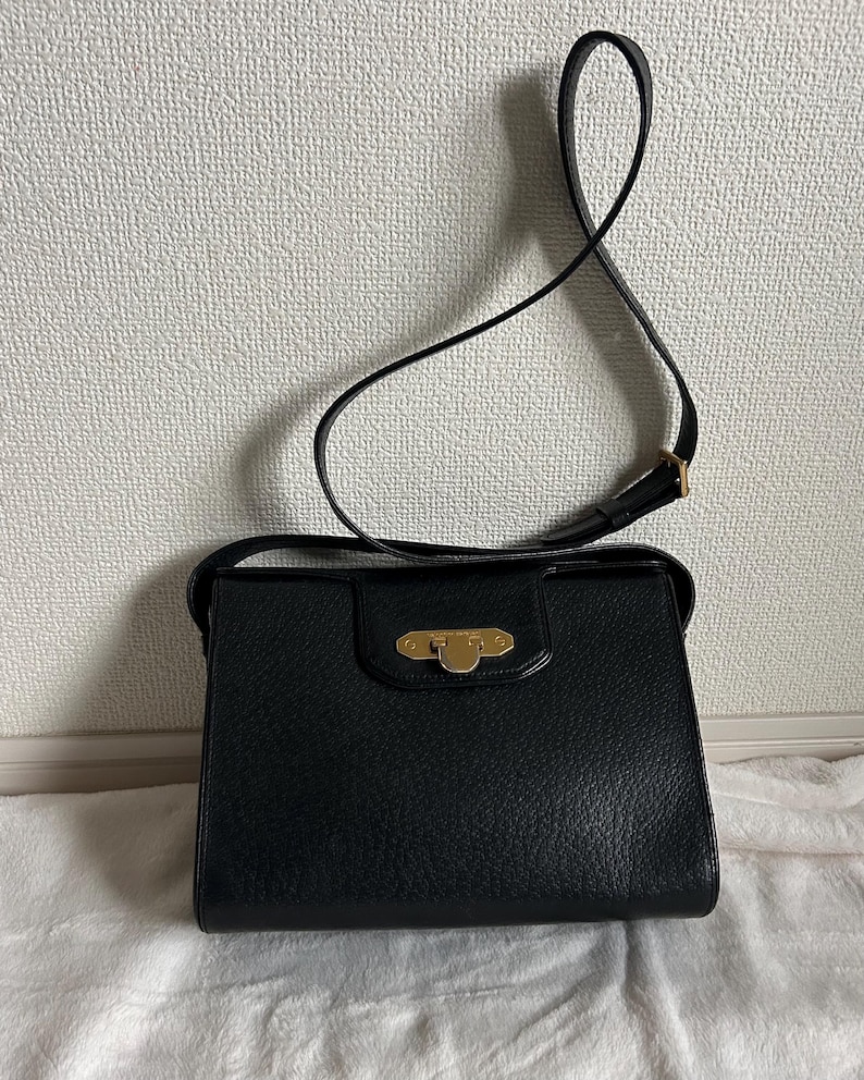 May include: Black leather shoulder bag with a rectangular shape and a long, thin strap. The bag features a gold-colored clasp on the front and a textured surface. The strap is adjustable and appears to be made of the same material as the bag.