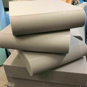 May include: A pile of light grey foam blocks, in various sizes and shapes. The foam has a smooth, slightly textured surface. The blocks appear freshly cut, with clean edges. The image suggests a material used for cushioning or padding, possibly for furniture.