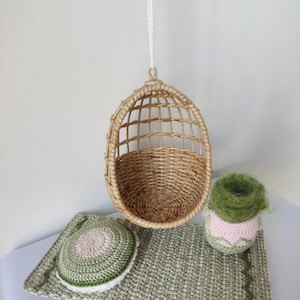May include: A miniature woven rattan hanging chair with a matching woven base, a crocheted green and white mat, and a crocheted pink and green plant pot. The items are arranged on a white surface.
