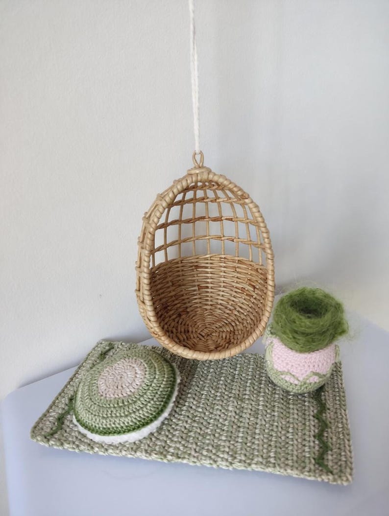 May include: A miniature woven rattan hanging chair with a matching woven base, a crocheted green and white mat, and a crocheted pink and green plant pot. The items are arranged on a white surface.