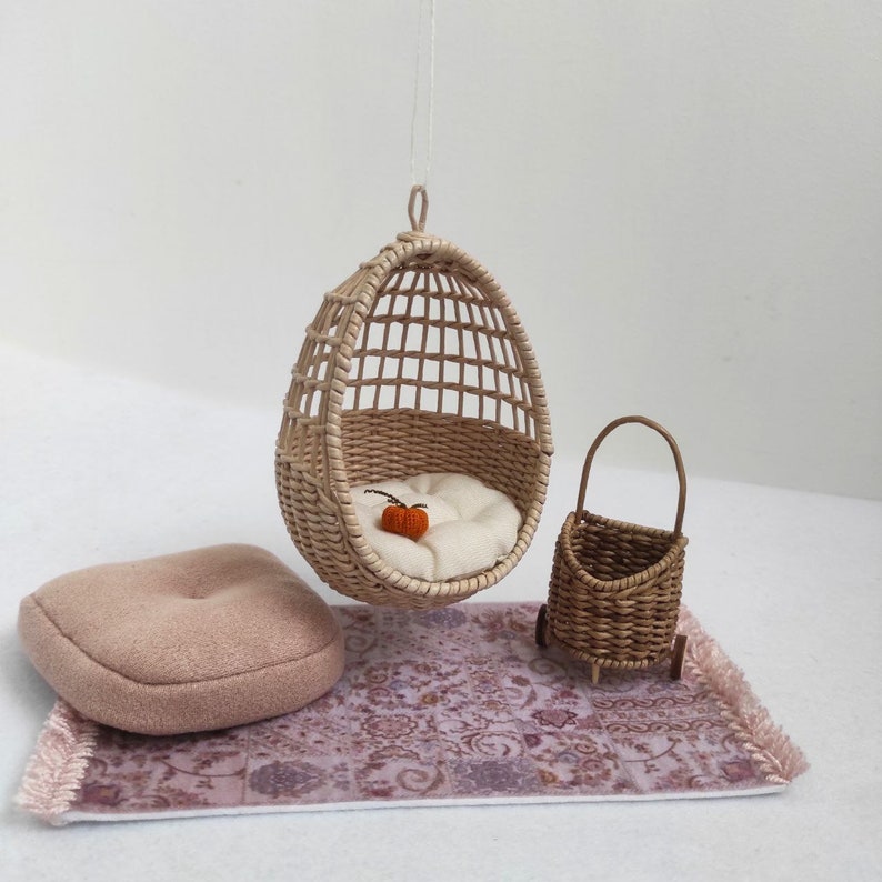May include: A miniature wicker hanging chair with a white cushion and a small orange pumpkin inside. A miniature wicker basket on wheels sits on a pink and purple patterned rug.