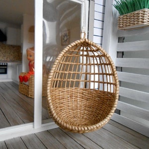 May include: A brown wicker hanging chair with a woven design. The chair is hanging from a white ceiling in a dollhouse.
