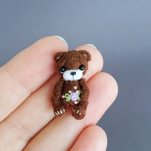 Op de afbeelding: Een kleine bruine gehaakte teddybeer met geborduurde bloemen op zijn borst. De beer heeft zwarte ogen en een witte neus.