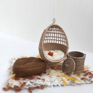 May include: A miniature wicker hanging chair with a white cushion and a small orange crocheted pumpkin. There is a brown leather ottoman and a wicker basket with a lid on a white and orange rug.