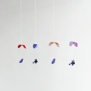 Könnte beinhalten: Vier bunte Hängegleiter mit Menschen, die daran hängen. Die Gleiter sind an Schnüren von der Decke abgehängt. Die Gleiter sind rot, blau, pink und lila.