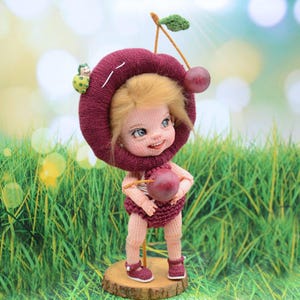 May include: A crocheted doll wearing a cherry-shaped hat and holding a cherry. The doll has blonde hair, blue eyes, and a smiling face. The doll is standing on a wooden base.
