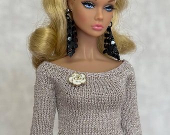 May include: A fashion doll with blonde hair styled in a half-up ponytail, blue eyes, and pink lips. The doll wears long black dangling earrings, a beige long-sleeved knit top with a metallic shimmer, a white flower brooch, and a black skirt.