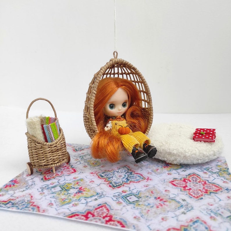 May include: A doll with red hair sits in a wicker hanging chair. The doll is wearing a yellow dress and black shoes. The chair is hanging over a white and blue patterned rug. There is a small wicker basket with colorful fabric rolls on wheels next to the rug.
