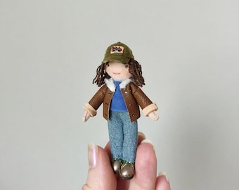 May include: A miniature doll with brown hair wearing an olive green baseball cap with a small red tractor emblem, a blue shirt, a brown faux leather jacket with white faux fur trim, blue denim-style pants, and small brown shoes.