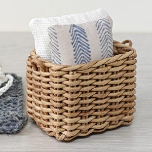 Op de afbeelding: Een kleine, geweven rieten mand gevuld met gevouwen witte en bedrukte stof met een blauw chevronontwerp. Een gebreide muts en een grijze gebreide vierkant liggen naast de mand.