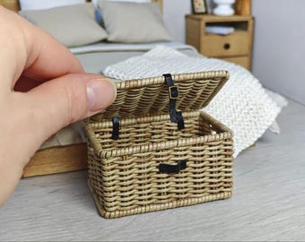 May include: A miniature rectangular woven wicker basket with a hinged lid. The basket features a light tan natural finish and is secured with a thin black faux leather strap and a small metal buckle closure on the front.