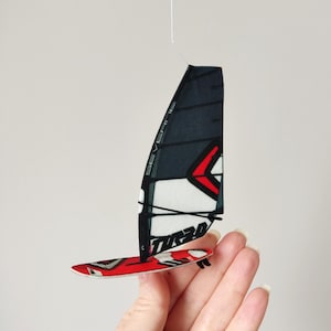 Puede incluir: Una vela y una tabla de windsurf en miniatura, con una vela negra y blanca y una tabla roja y blanca. La vela tiene la palabra "SEVEN" impresa y la tabla tiene la palabra "TOR20" impresa.