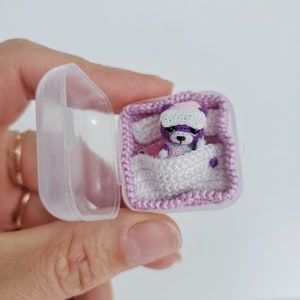May include: A small purple crocheted teddy bear wearing a white crocheted hat is lying in a pink crocheted bed inside a clear plastic container.