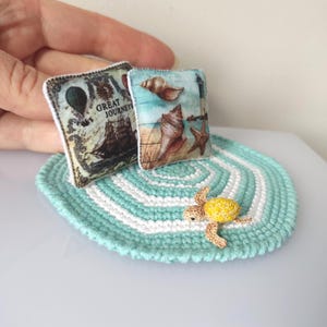 Può includere: Set in miniatura a tema nautico con un tappeto all'uncinetto turchese e bianco, due piccoli cuscini con stampe di conchiglie e navi e una statuina di tartaruga gialla e marrone. I cuscini hanno un bordo bianco e il tappeto è rotondo.