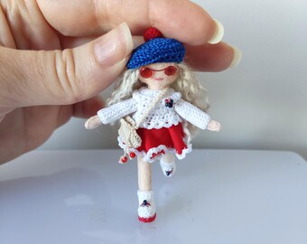 May include: A small crocheted doll wearing a blue beret with a red pom-pom, red sunglasses, a white crocheted sweater, a red skirt with white trim, and white crocheted shoes with red accents.