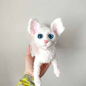 Può includere: Un gatto di peluche bianco con grandi occhi blu, orecchie rosa e naso rosa. Il gatto ha una consistenza soffice e un delicato fiocco rosa intorno al collo. È un articolo fatto a mano.