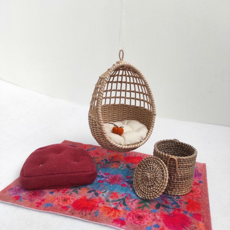 May include: A miniature wicker hanging chair with a white cushion and a small orange pumpkin inside. The chair is on a colorful floral rug with a red cushion and a wicker basket with a lid.
