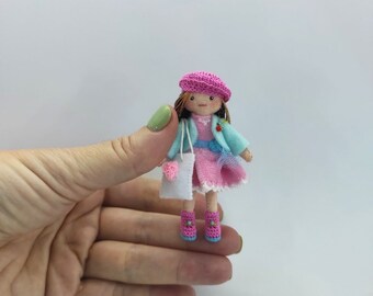 May include: A small crocheted doll wearing a pink dress, blue jacket, and pink hat. The doll is holding a white crocheted bag and is wearing pink shoes.