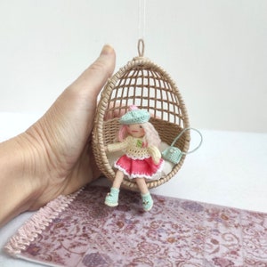 May include: A small doll wearing a pink and white dress, a light blue hat, and holding a small light blue purse sits in a hanging wicker chair. The chair is suspended from the ceiling and the doll is looking to the right. The doll is sitting on a pink and white patterned rug.