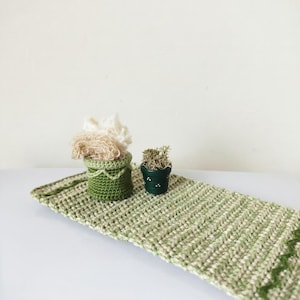 Puede incluir: Camino de mesa de ganchillo verde y beige hecho a mano con un patrón en zigzag en un extremo. Una pequeña cesta de ganchillo verde y una pequeña maceta verde con una planta se encuentran sobre el camino de mesa. La cesta contiene hilo beige.