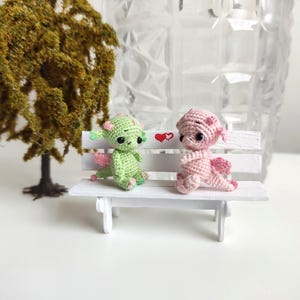 May include: Two small crocheted dragons sitting on a white bench. The dragon on the left is green and the dragon on the right is pink. There are two red hearts painted on the bench behind the dragons.