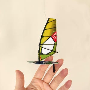 Puede incluir: Un windsurfista en miniatura con una vela amarilla y verde, una sección de vela blanca y una tabla azul y verde. La vela tiene un logotipo rojo. El windsurfista está suspendido por un fino hilo blanco.