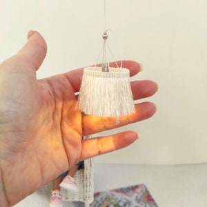 Puede incluir: Pantalla de lámpara en miniatura con flecos de color crema. La pequeña pantalla se sostiene en una mano, suspendida por un alambre fino. La pantalla tiene una parte superior plateada y flecos. Diseñada para una casa de muñecas.