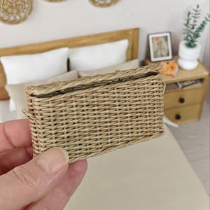 May include: A small, rectangular woven basket in a light brown color. The basket is held in a hand and appears to be a miniature or dollhouse accessory. The basket has a lid and is made of natural fibers.