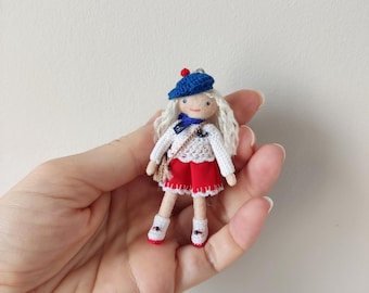 May include: A handmade miniature doll with white hair, wearing a blue beret with a red pom-pom, a white sweater, red shorts, and white shoes. The doll has a small brown purse and a blue scarf. The doll is held in a person's hand.