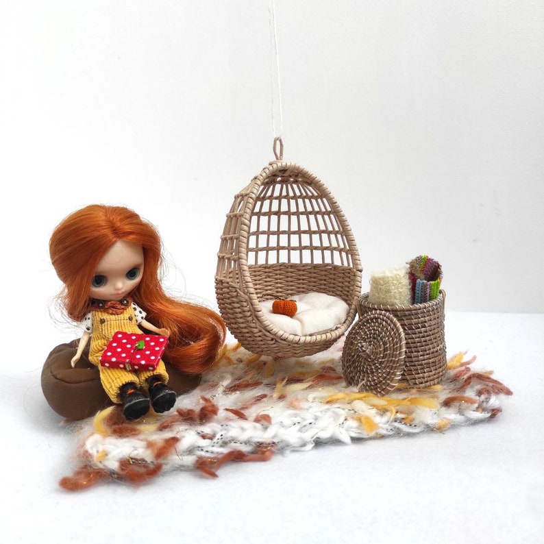 May include: A doll with red hair sits on a brown cushion with a red and white patterned book. A wicker hanging chair with a white cushion hangs above a white and brown rug. A wicker basket with a lid sits on the rug.
