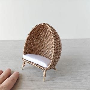 Könnte beinhalten: Ein Miniatur-Korbstuhl mit einem weißen Kissen. Der Stuhl ist kuppelförmig und hat eine abgerundete Rückenlehne.