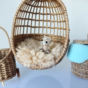 May include: A small crocheted bear sits in a wicker egg-shaped chair. The chair is filled with white fluffy material. A wicker basket with a blue fabric liner sits on a white surface.