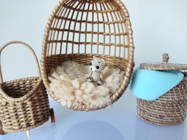 May include: A small crocheted bear sits in a wicker egg-shaped chair. The chair is filled with white fluffy material. A wicker basket with a blue fabric liner sits on a white surface.