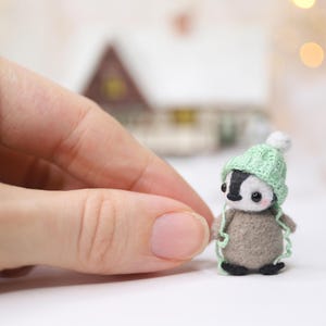 May include: A miniature felt penguin figurine wearing a light green knitted hat with a white pom-pom. The penguin has a black and white face and a grey body. The background is blurred, with a small house and bokeh lights.