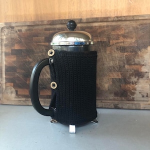 May include: A black knitted cosy for a French press coffee maker. The cosy has a silver metal base and a glass carafe with a black lid.