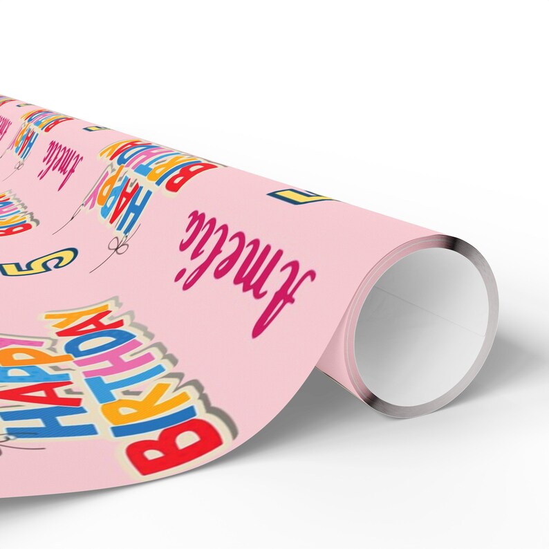 May include: A roll of pink wrapping paper featuring the words "HAPPY BIRTHDAY" in vibrant colours and the name "Amelia". The text is in various colours, including red, blue, yellow, and orange. The paper is rolled up.