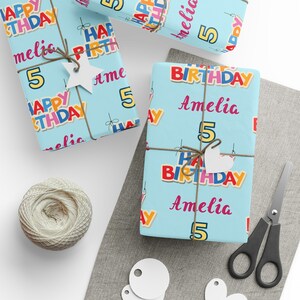 May include: Gifts wrapped in light blue birthday paper, patterned with colourful "HAPPY BIRTHDAY" text, the name "Amelia", and the number "5". The presents are tied with twine and decorated with white tags. Scissors and a ball of twine are also visible.