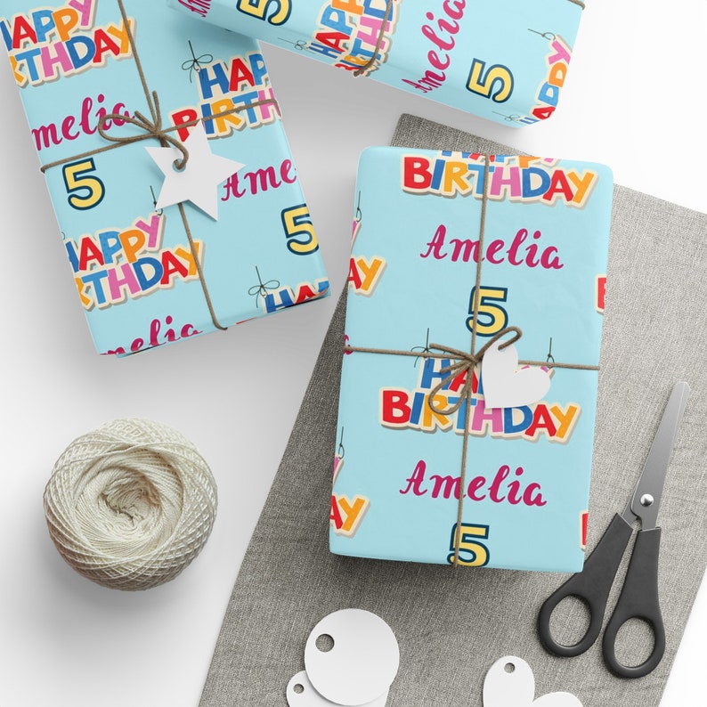 May include: Gifts wrapped in light blue birthday paper, patterned with colourful "HAPPY BIRTHDAY" text, the name "Amelia", and the number "5". The presents are tied with twine and decorated with white tags. Scissors and a ball of twine are also visible.