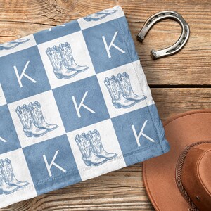 Peut inclure: Une couverture pour bébé à carreaux bleus et blancs avec un motif répété de bottes de cowboy et de la lettre "K".